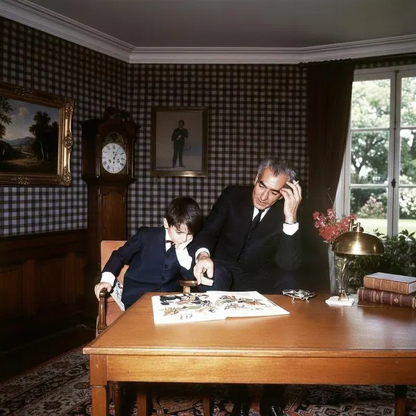 An adult man and a young boy in dark suits look at a book on a wooden table — a public domain photograph from The Shah of Iran archive documenting the Pahlavi era.