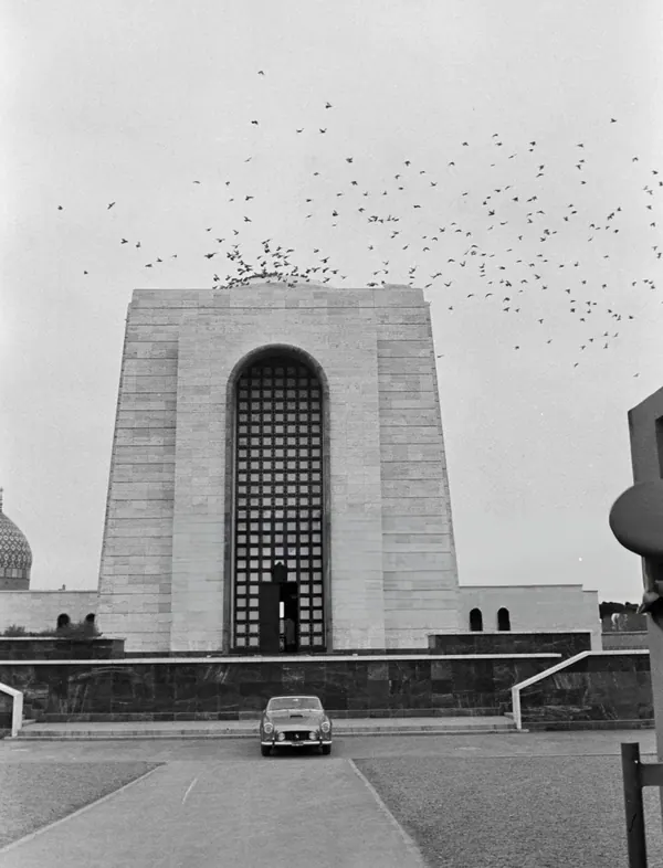 A car sits in front of a large stone monument with a high arched window and flying birds — a public domain photograph from The Shah of Iran archive documenting the Pahlavi era.