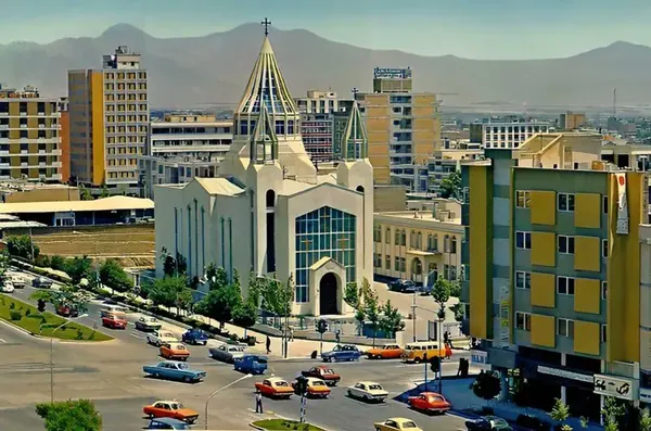 A church with a pointed spire stands among multi-story buildings and a street with parked cars — a public domain photograph from The Shah of Iran archive documenting the Pahlavi era.
