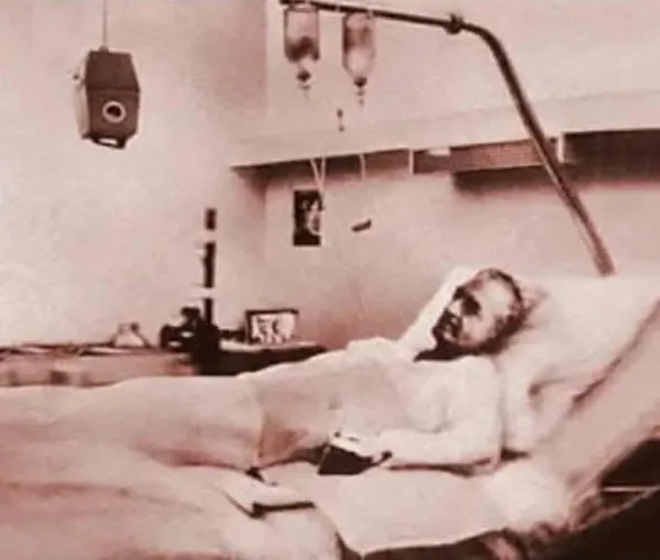 An elderly man lies in a hospital bed with medical equipment and IV bags hanging above him — a public domain photograph from The Shah of Iran archive documenting the Pahlavi era.