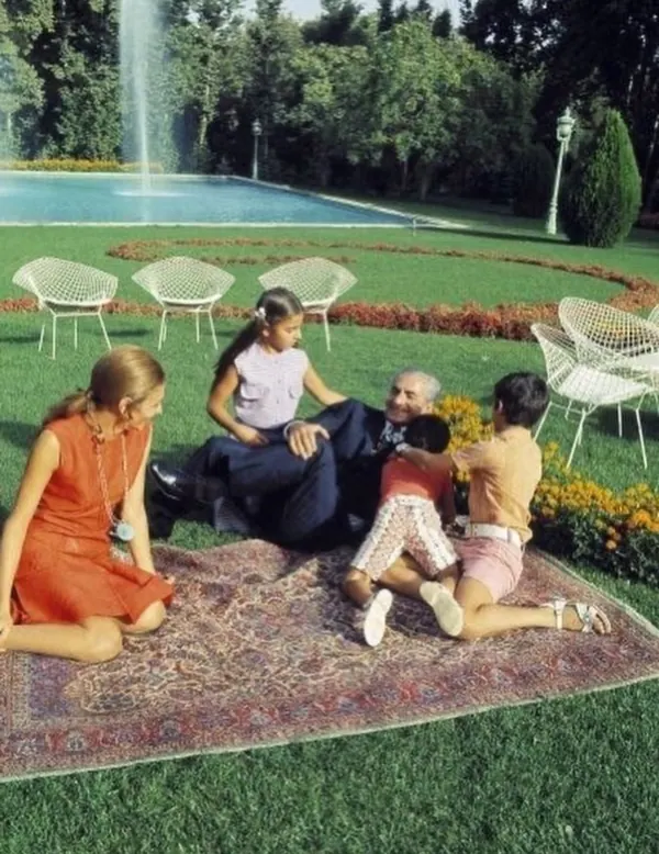 A family sits on a rug in a garden with a fountain and white chairs in the background — a public domain photograph from The Shah of Iran archive documenting the Pahlavi era.