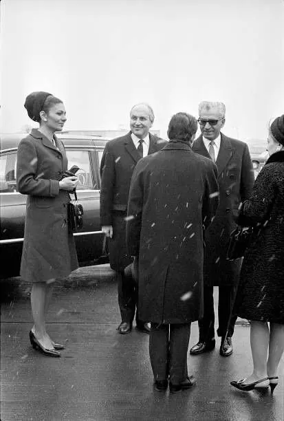 Four adults in formal coats and hats stand outdoors near a dark car on a wet surface — a public domain photograph from The Shah of Iran archive documenting the Pahlavi era.