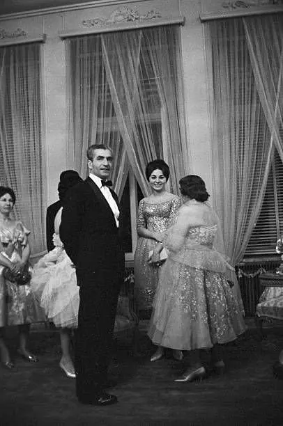 Four adults in formal evening attire stand and talk in a room with large curtained windows — a public domain photograph from The Shah of Iran archive documenting the Pahlavi era.