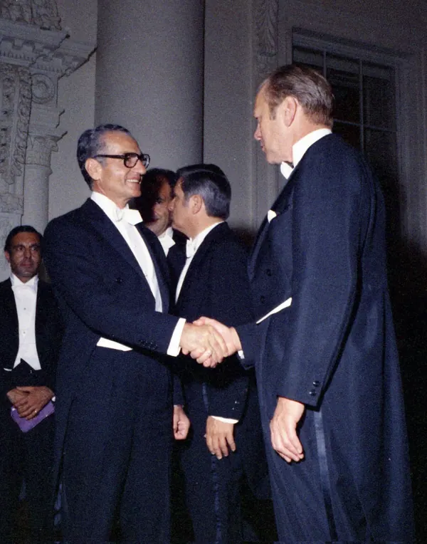 Four men in formal black tuxedos stand together, with two men shaking hands in the foreground — a public domain photograph from The Shah of Iran archive documenting the Pahlavi era.