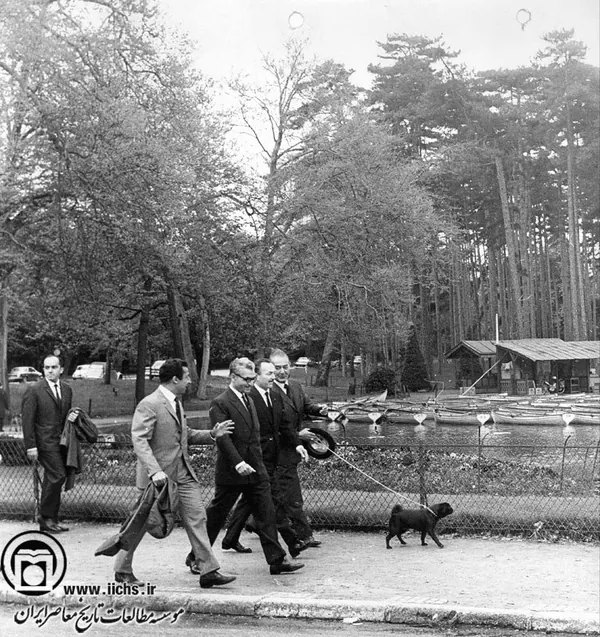 Four men in suits walk along a path near a lake with boats and a small dog — a public domain photograph from The Shah of Iran archive documenting the Pahlavi era.