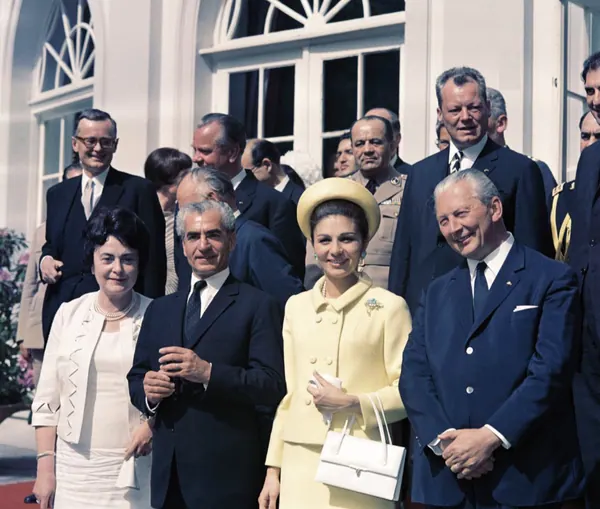 A group of men and women in formal attire stand together in front of a white building — a public domain photograph from The Shah of Iran archive documenting the Pahlavi era.
