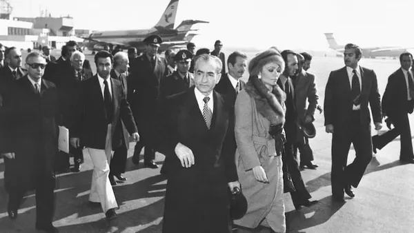 A group of men in suits and Farah Pahlavi, Empress of Iran, in a fur-trimmed coat walk across an airfield — a public domain photograph from The Shah of Iran archive documenting the Pahlavi era.