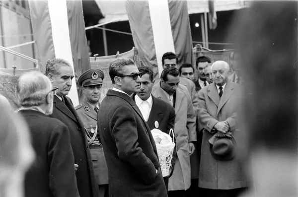A group of men in suits and one in military uniform stand together outdoors near large flags — a public domain photograph from The Shah of Iran archive documenting the Pahlavi era.