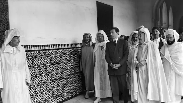 A group of men in traditional white robes and one man in a suit walk through a hallway — a public domain photograph from The Shah of Iran archive documenting the Pahlavi era.
