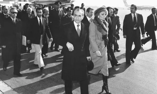 A group of people in formal attire walk across a paved airfield with an airplane in the background — a public domain photograph from The Shah of Iran archive documenting the Pahlavi era.