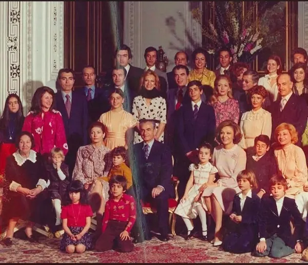 A large group of adults and children in formal attire pose for a portrait in a decorated room — a public domain photograph from The Shah of Iran archive documenting the Pahlavi era.
