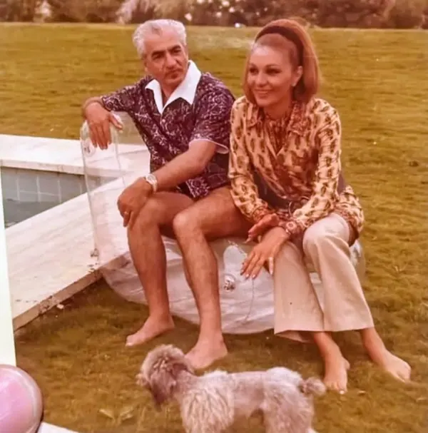 His Majesty, the Shah of Iran and woman sit outdoors on a clear chair with a small dog on the grass — a public domain photograph from The Shah of Iran archive documenting the Pahlavi era.