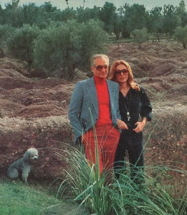 His Majesty, the Shah of Iran in a blue blazer and red pants stands with Farah Pahlavi, Empress of Iran, in black attire outdoors — a public domain photograph from The Shah of Iran archive documenting the Pahlavi era.