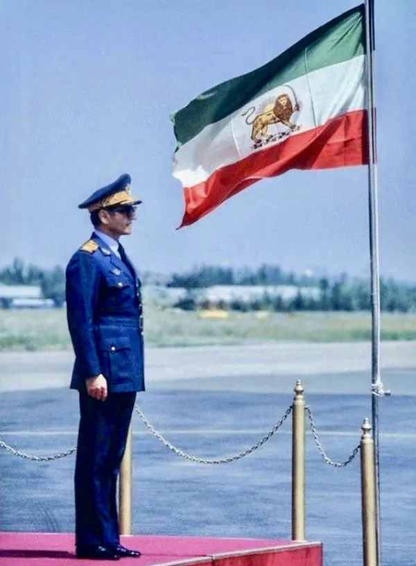 His Majesty, the Shah of Iran in a blue military uniform stands at attention beside a flagpole with a waving flag — a public domain photograph from The Shah of Iran archive documenting the Pahlavi era.