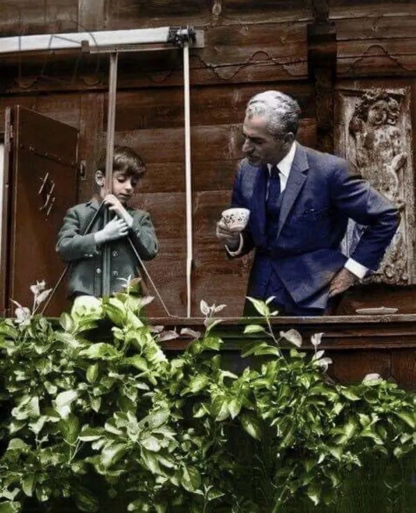 His Majesty, the Shah of Iran in a blue suit holds a cup while standing beside a young boy in a jacket — a public domain photograph from The Shah of Iran archive documenting the Pahlavi era.