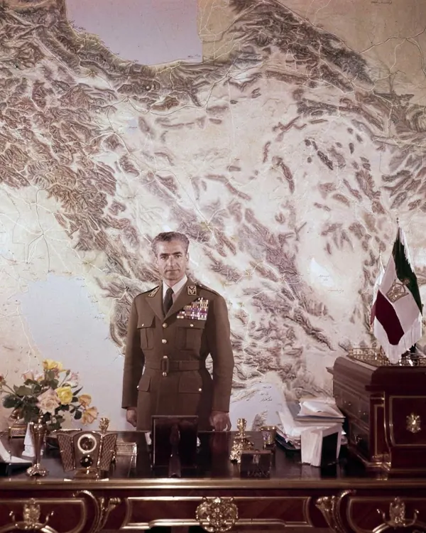 His Majesty, the Shah of Iran in a brown military uniform stands before a large map and a wooden desk — a public domain photograph from The Shah of Iran archive documenting the Pahlavi era.