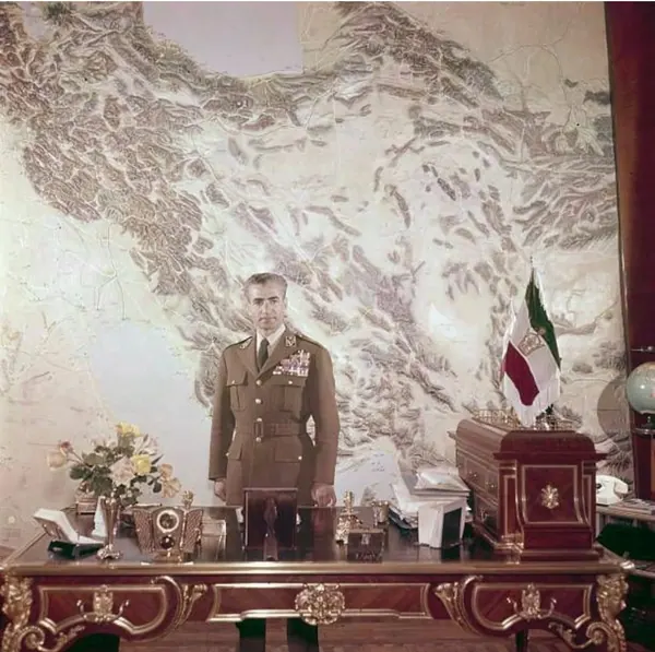 His Majesty, the Shah of Iran in a brown military uniform stands behind an ornate desk in front of a large map — a public domain photograph from The Shah of Iran archive documenting the Pahlavi era.