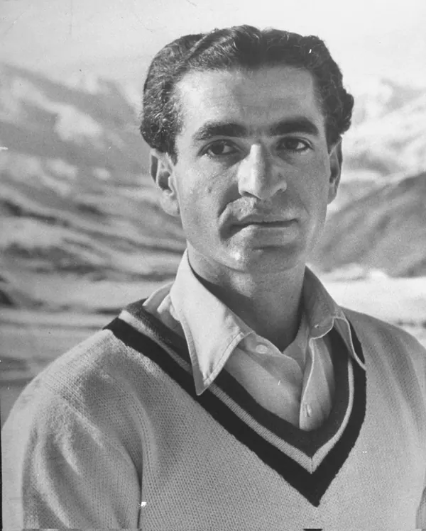 His Majesty, the Shah of Iran in a collared shirt and V-neck sweater poses against a backdrop of snowy mountains — a public domain photograph from The Shah of Iran archive documenting the Pahlavi era.