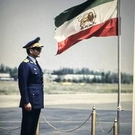 His Majesty, the Shah of Iran in a dark blue military uniform stands at attention beside a waving tricolor flag — a public domain photograph from The Shah of Iran archive documenting the Pahlavi era.
