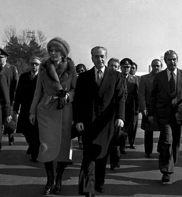 His Majesty, the Shah of Iran in a dark coat and Farah Pahlavi, Empress of Iran, in a fur-trimmed coat walk together outdoors with attendants — a public domain photograph from The Shah of Iran archive documenting the Pahlavi era.