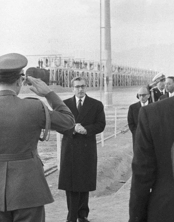 His Majesty, the Shah of Iran in a dark coat and glasses stands while a uniformed officer salutes him from the side — a public domain photograph from The Shah of Iran archive documenting the Pahlavi era.