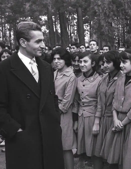 His Majesty, the Shah of Iran in a dark coat stands before a group of young people in light-colored uniforms in woods — a public domain photograph from The Shah of Iran archive documenting the Pahlavi era.