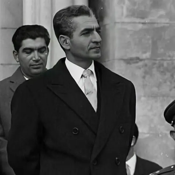 His Majesty, the Shah of Iran in a dark double-breasted coat and tie stands beside another man in a suit — a public domain photograph from The Shah of Iran archive documenting the Pahlavi era.