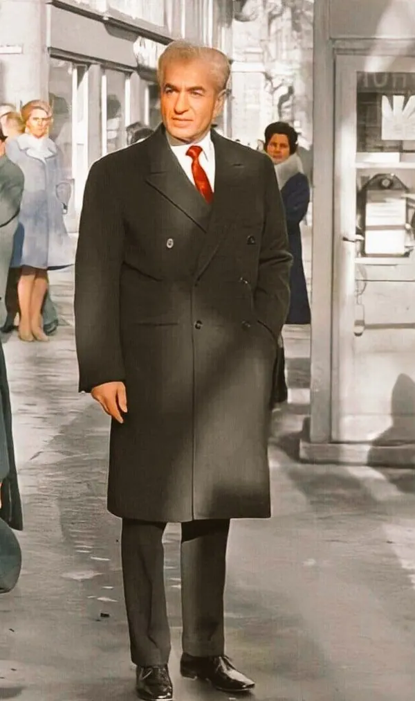 His Majesty, the Shah of Iran in a dark double-breasted overcoat and red tie walks along a city sidewalk near a phone booth — a public domain photograph from The Shah of Iran archive documenting the Pahlavi era.