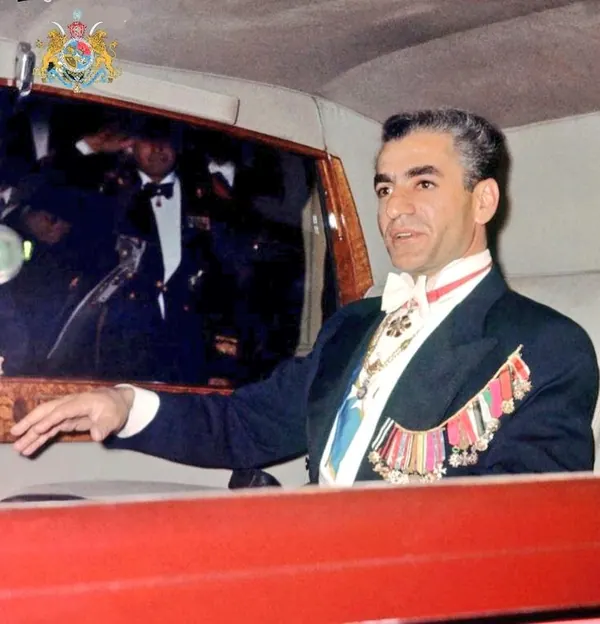 His Majesty, the Shah of Iran in a dark formal suit with medals and a white bow tie sits in a vehicle — a public domain photograph from The Shah of Iran archive documenting the Pahlavi era.