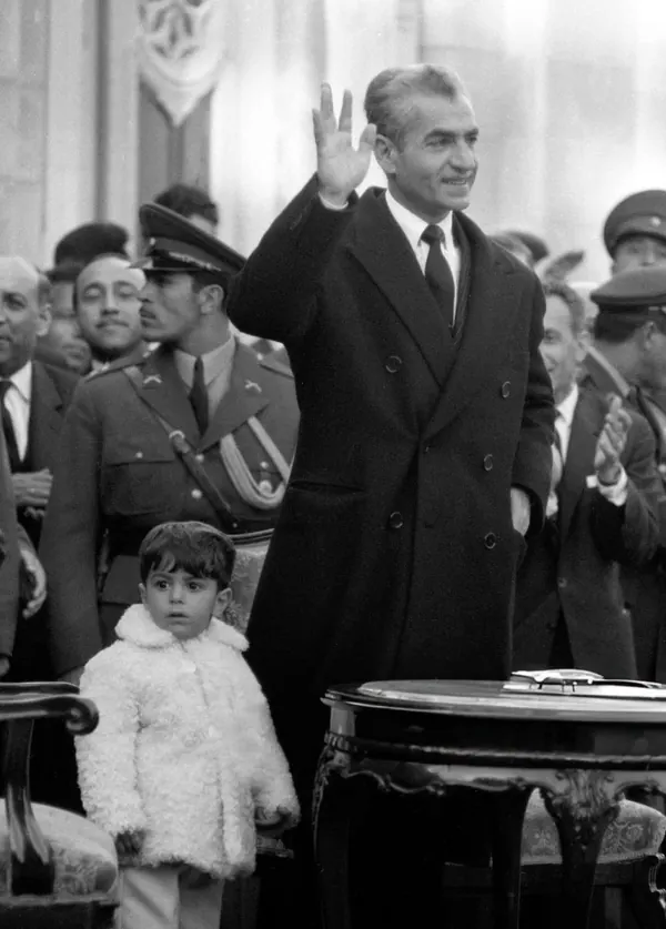 His Majesty, the Shah of Iran in a dark overcoat waves his hand while standing beside a small child in a white coat — a public domain photograph from The Shah of Iran archive documenting the Pahlavi era.