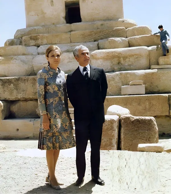 His Majesty, the Shah of Iran in a dark suit and Farah Pahlavi, Empress of Iran, in a patterned dress stand before stone ruins — a public domain photograph from The Shah of Iran archive documenting the Pahlavi era.