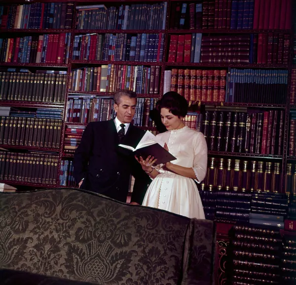 His Majesty, the Shah of Iran in a dark suit and Farah Pahlavi, Empress of Iran, in a white dress read a book together in a library — a public domain photograph from The Shah of Iran archive documenting the Pahlavi era.