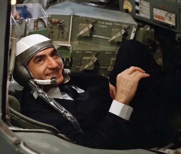 His Majesty, the Shah of Iran in a dark suit and flight helmet reclines inside a cramped cockpit with technical equipment — a public domain photograph from The Shah of Iran archive documenting the Pahlavi era.