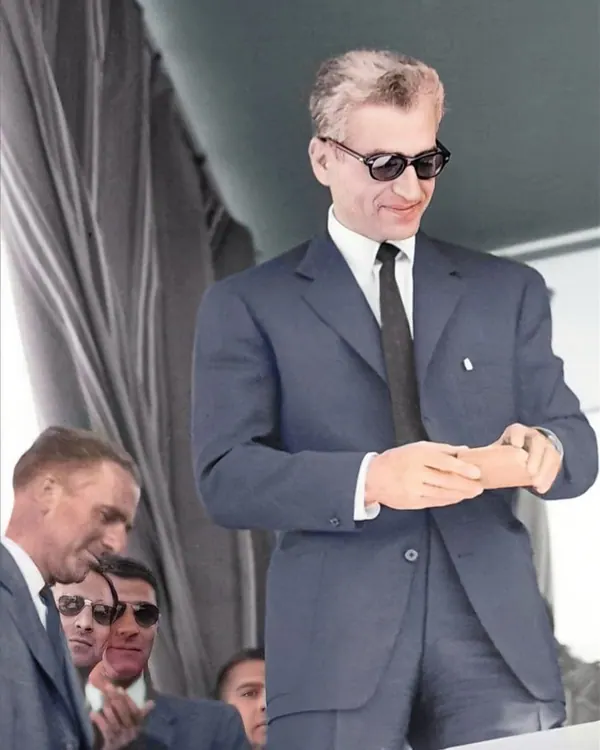 His Majesty, the Shah of Iran in a dark suit and sunglasses looks down while adjusting his cuff — a public domain photograph from The Shah of Iran archive documenting the Pahlavi era.