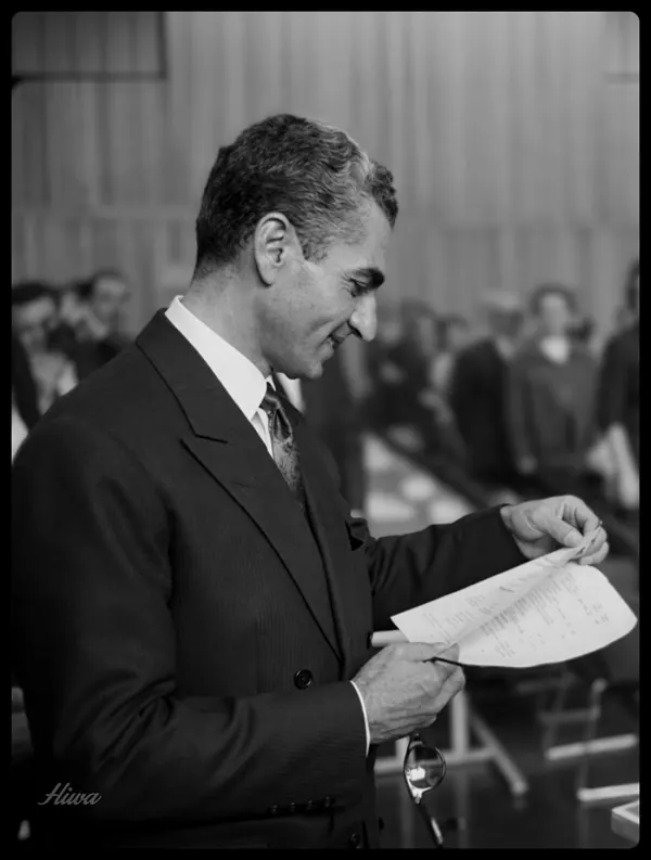 His Majesty, the Shah of Iran in a dark suit and tie stands in profile reading a piece of paper — a public domain photograph from The Shah of Iran archive documenting the Pahlavi era.