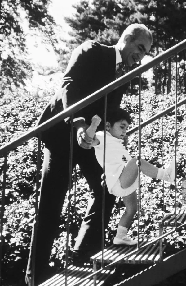 His Majesty, the Shah of Iran in a dark suit helps a small child in white clothing walk down metal stairs — a public domain photograph from The Shah of Iran archive documenting the Pahlavi era.