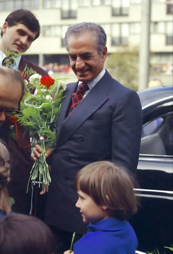 His Majesty, the Shah of Iran in a dark suit holds a bouquet of flowers while standing beside a black car — a public domain photograph from The Shah of Iran archive documenting the Pahlavi era.