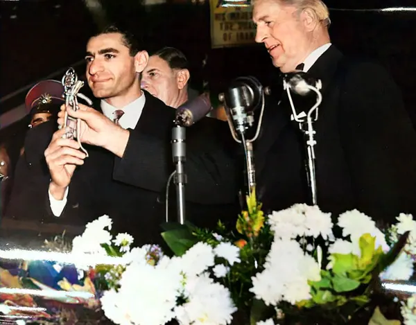 His Majesty, the Shah of Iran in a dark suit holds a small trophy while standing behind microphones and white flowers — a public domain photograph from The Shah of Iran archive documenting the Pahlavi era.