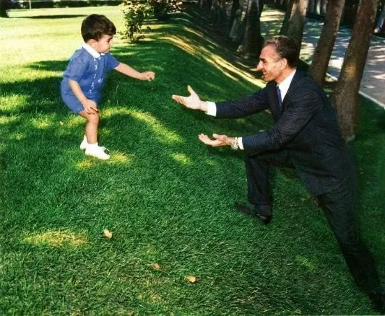 His Majesty, the Shah of Iran in a dark suit reaches out to a young child in blue clothing on a grassy lawn — a public domain photograph from The Shah of Iran archive documenting the Pahlavi era.