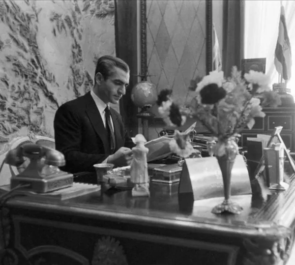 His Majesty, the Shah of Iran in a dark suit sits at a desk with a telephone, globe, and flowers — a public domain photograph from The Shah of Iran archive documenting the Pahlavi era.