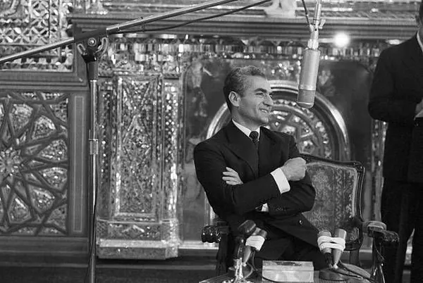 His Majesty, the Shah of Iran in a dark suit sits in an ornate chair with arms crossed near a microphone — a public domain photograph from The Shah of Iran archive documenting the Pahlavi era.