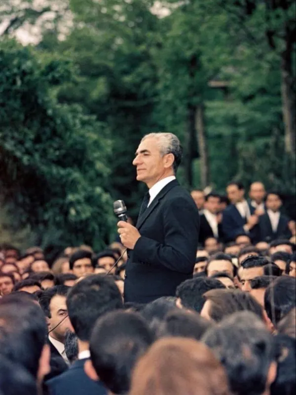 His Majesty, the Shah of Iran in a dark suit speaks into a microphone before a large crowd of people outdoors — a public domain photograph from The Shah of Iran archive documenting the Pahlavi era.