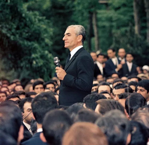 His Majesty, the Shah of Iran in a dark suit speaks into a microphone before a dense crowd of people outdoors — a public domain photograph from The Shah of Iran archive documenting the Pahlavi era.