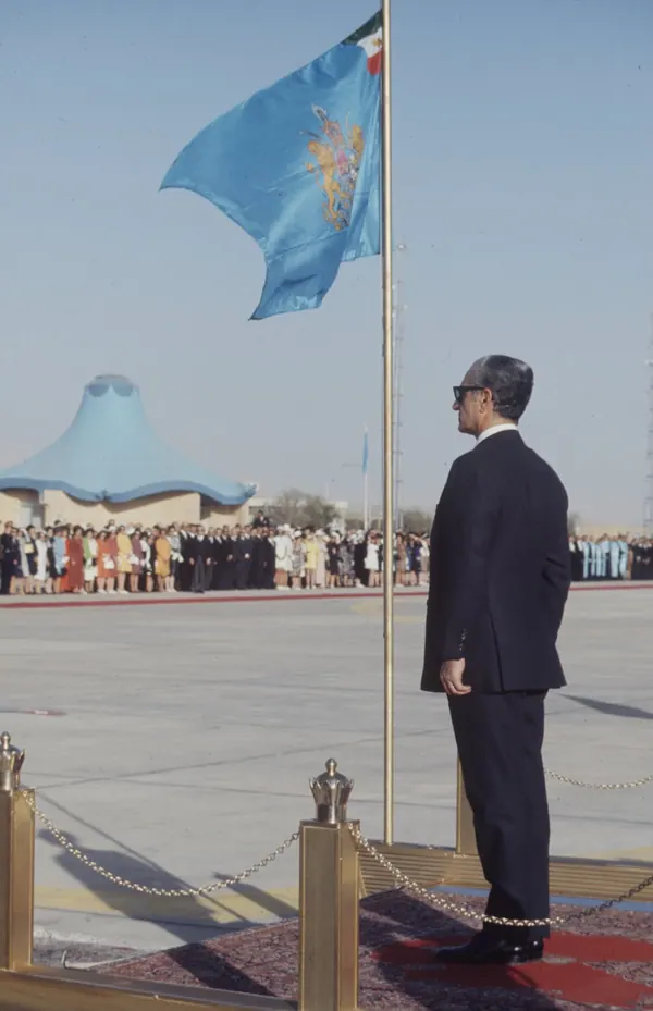 His Majesty, the Shah of Iran in a dark suit stands at attention beside a flagpole with a blue flag — a public domain photograph from The Shah of Iran archive documenting the Pahlavi era.