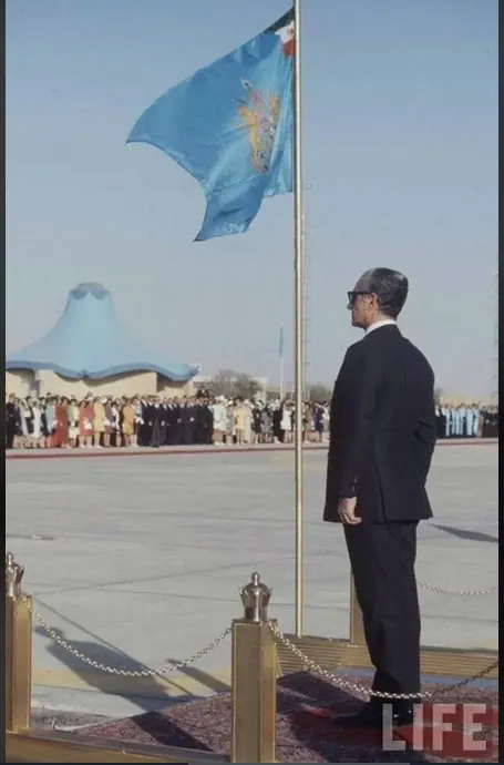 His Majesty, the Shah of Iran in a dark suit stands beside a flagpole while a crowd watches in the background — a public domain photograph from The Shah of Iran archive documenting the Pahlavi era.