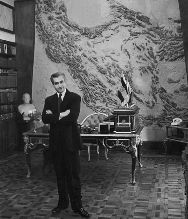 His Majesty, the Shah of Iran in a dark suit stands with arms crossed in a room with a large relief map — a public domain photograph from The Shah of Iran archive documenting the Pahlavi era.
