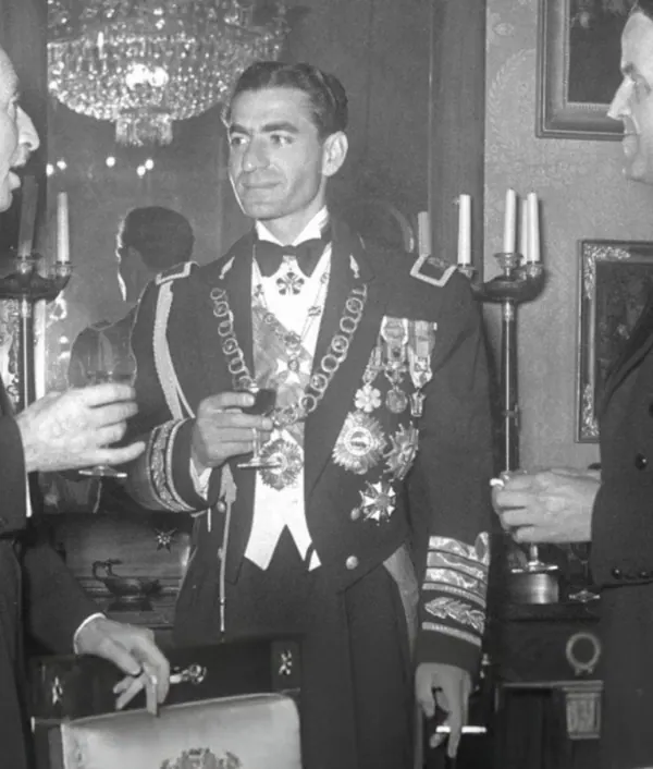 His Majesty, the Shah of Iran in a decorated military uniform holds a glass of wine while standing in a room — a public domain photograph from The Shah of Iran archive documenting the Pahlavi era.