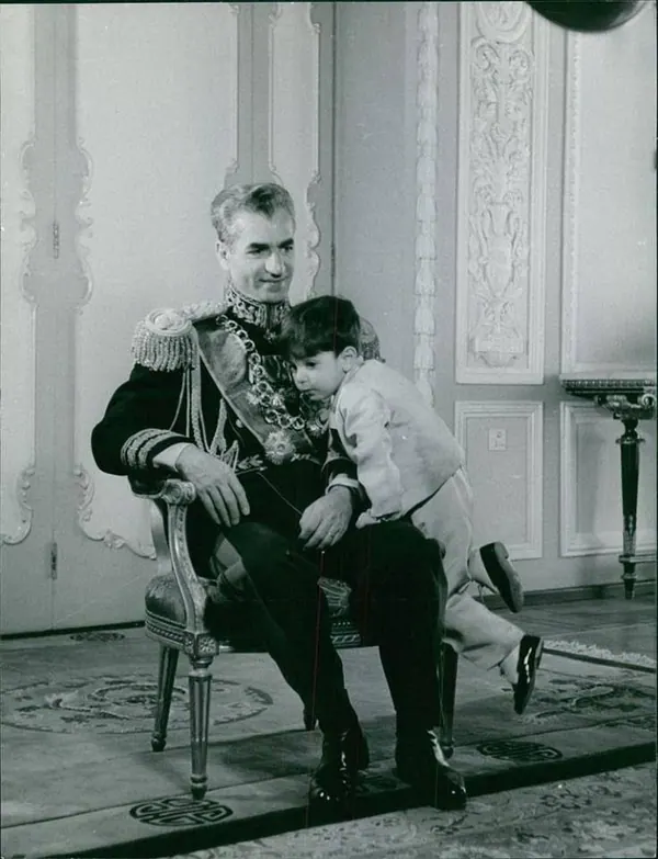 His Majesty, the Shah of Iran in a decorated military uniform sits in a chair holding a young child in light clothing — a public domain photograph from The Shah of Iran archive documenting the Pahlavi era.