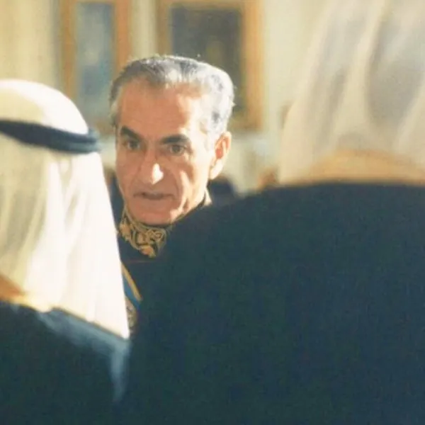 His Majesty, the Shah of Iran in a decorated uniform looks toward others wearing traditional white head coverings in an indoor setting — a public domain photograph from The Shah of Iran archive documenting the Pahlavi era.