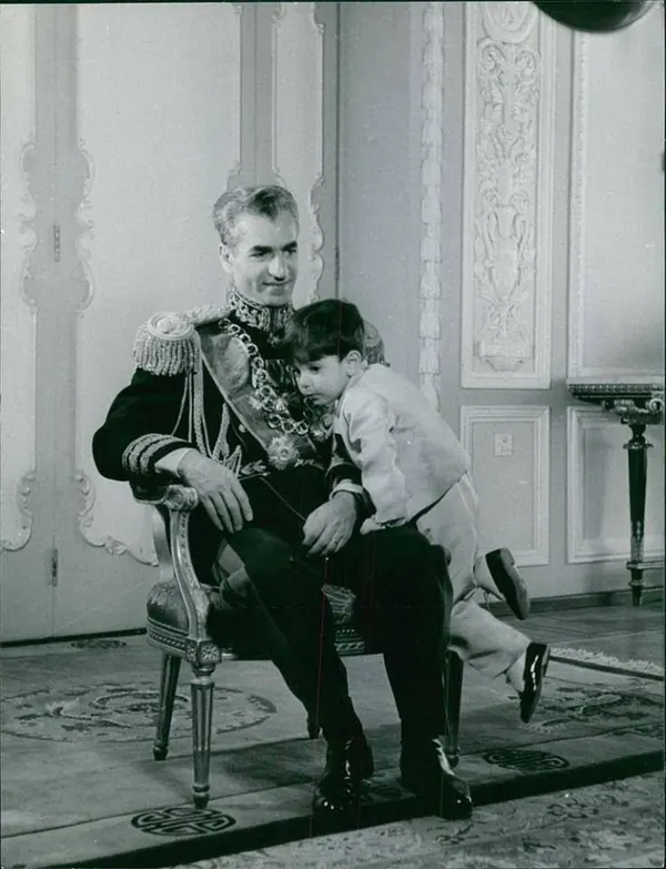 His Majesty, the Shah of Iran in a formal military uniform sits in a chair while a small child leans against him — a public domain photograph from The Shah of Iran archive documenting the Pahlavi era.